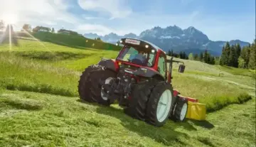  Ein Traktor mit Anhänger steht auf einem grünen Hügel, umgeben von saftigem Gras und klarem Himmel.