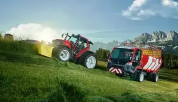  Ein Traktor mit Anhänger steht auf einem grünen Hügel, umgeben von saftigem Gras und klarem Himmel.